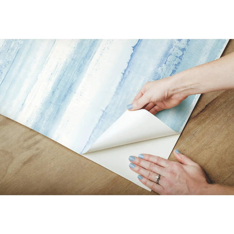 Hands peeling Tan Watercolor Stripe Peel And Stick Wallpaper Roll 20.5 Inch Wide By 16.5 Foot Long for easy DIY installation on wood.