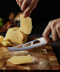 Five Inch Serrated Cheese Knife Crafted With Premium Steel Core And A Super Slick Hollow Blade For Butter Smooth Cuts That Slide Off Easy Plus A Gotta Have Fork Tip For Zero Mess Party Spread cutting cheese on a wooden board.