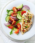 Grilled chicken breast with a side of fresh tomato, cucumber, red onion, and olive salad on a white plate.