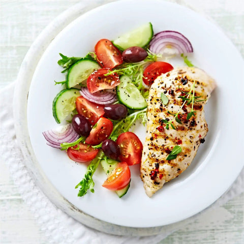 Grilled chicken breast with a side of fresh tomato, cucumber, red onion, and olive salad on a white plate.