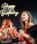 People celebrating at a party under 'Happy Birthday' neon sign, showcasing Smile Face Neon Sign Birthday Light 13 By 13 Inches Portable USB Powered Warm White Glow For Parties Take Anywhere Easy To Install With 2 Meter USB Cable Photo Backdrop And Party Ready Decor For Events.