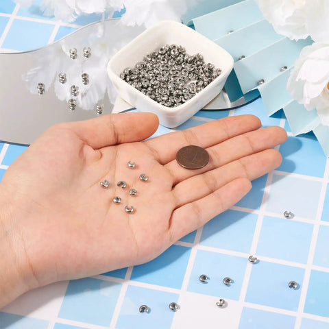 Hand holding stainless steel crimp bead covers 6x5x3mm, with bowl of 200 pieces for jewelry making background.