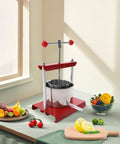 Heavy Duty T Handle Fruit Wine Press With Dual Stainless Steel Barrels on kitchen table with fruits and vegetables.