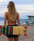 Arbor longboard with striped wooden deck and orange wheels held by woman on the beach