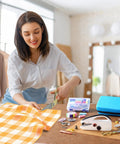 Woman using Heavy Duty Handheld Sewing Machine Kit With Blue Leather Storage Case And 110 Piece Sewing Kit, fabrics and sewing tools displayed.