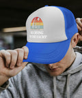 Man wearing a blue and white trucker hat with 'No Crying on the Yacht' text, adjusting brim, casual hoodie outfit