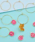 Rose Red Enamel Alphabet Charms And Golden Wine Glass Marker Rings Set on a blue background with colorful charms visible