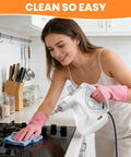 Powerful Portable Handheld Steamer Shooting Scalding Vapor To Wreck Stubborn Pan Grease And Baked On Filth On Tile Linoleum And Fabric Seats With 14 Heavy Duty Tools For No Sweat Scrubbing And Spotless Shine being used by a woman on a stovetop.