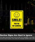 Two Pack Smile You're On Camera Video Surveillance Sign For Home And Business 10x7 aluminum sign on a fence at night.