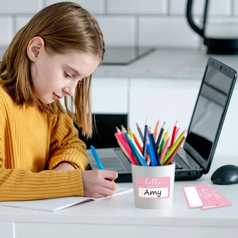 Child writing at desk with Light Pink Hello My Name Is Stickers Set, 300 Pcs Name Tag Labels On A Roll nearby