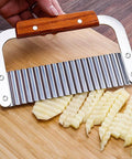 Wooden-handle stainless steel crinkle cutter with a wavy blade slicing crinkle-cut potatoes on a cutting board