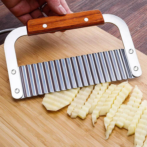 Wooden-handle stainless steel crinkle cutter with a wavy blade slicing crinkle-cut potatoes on a cutting board