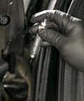ARES 70004 precision oiler pen applicator held by gloved hand near a car door, used to apply lubricant in tight spaces.