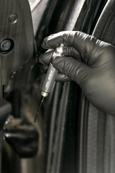ARES 70004 precision oiler pen applicator held by gloved hand near a car door, used to apply lubricant in tight spaces.