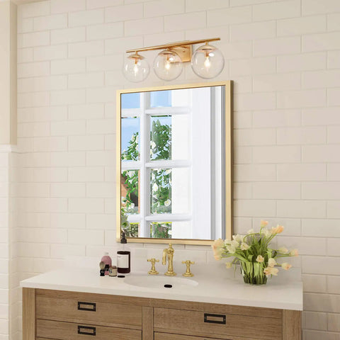 Three Light Gold Vanity Light Fixture With Clear Globe Glass Shades And Slim Taper Arm For Modern Bathroom Decor Exposed Bulbs And Bright Even Illumination With Sleek Gold Finish above mirror.