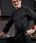 Heavy Duty Chef Coat With Chest And Sleeve Pocket Plus Hella Breathable Fabric That Keeps You Cool In The Heat Top Drawer Restaurant Cook Uniform worn by a chef in a kitchen setting.