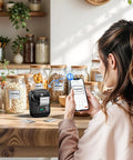 Woman using Bluetooth Portable Label Printer For Small Biz Fast Thermal Label Maker For Barcodes Addresses Clothing Pricing Mail Jewelry Cables And More Works With Phone And PC to label kitchen jars.