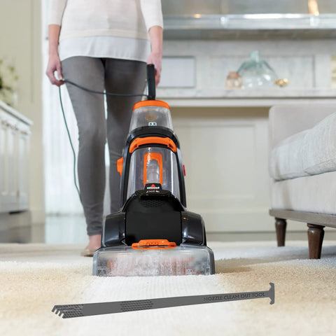 Front view of an orange-and-black carpet cleaner with a transparent water tank; nozzle clean-out tool on the carpet