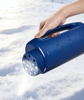 Person using handheld seed shaker and spreader with 90 oz capacity, blue, on snow for ice melt application.