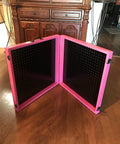 Pink foldable pegboard standing on wooden floor in front of cabinet, featuring black peg panels and corner hinges for stability.