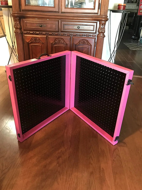 Pink foldable pegboard standing on wooden floor in front of cabinet, featuring black peg panels and corner hinges for stability.