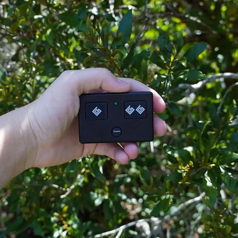 Hand holding AXS1 3-Button Gate Opener Remote Transmitter for Automatic Gates, Authentic OEM, easy pairing, in garden setting.
