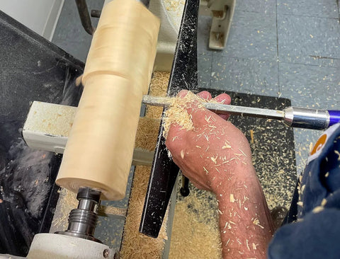 Close-up of a carbide wood-turning tool with a color-coded 12-inch aluminum lathe handle shaping a light wood blank on a lathe.