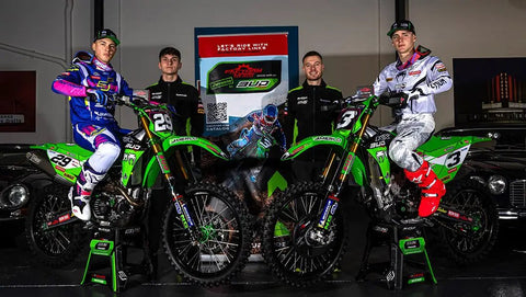 Two motocross riders on green bikes with two team members standing behind, all in racing gear and uniforms.