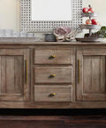 Wooden sideboard with antique brass finger pulls, three drawers and two cabinet doors, styled with decor and glassware.