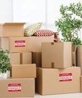 Boxes with "Please Handle With Care Fragile" stickers in a room, displaying "Fragile Stickers 2x3 Inches For Shipping And Moving With Permanent Adhesive 500 Labels On One Roll Please Handle With Care Do Not Drop Warning Labels".