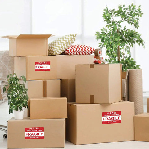 Boxes with "Please Handle With Care Fragile" stickers in a room, displaying "Fragile Stickers 2x3 Inches For Shipping And Moving With Permanent Adhesive 500 Labels On One Roll Please Handle With Care Do Not Drop Warning Labels".