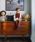 Child sitting on a mid-century modern cabinet with toys and decor items, including a vintage typewriter and a lamp with a lotus shade.
