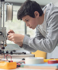Person working on a 3D printer, adjusting components, surrounded by tools and materials on a table in a workshop setting.
