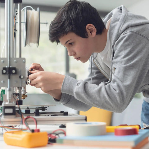 Person working on a 3D printer, adjusting components, surrounded by tools and materials on a table in a workshop setting.