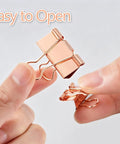 Close-up of hands holding two Rose Gold Binder Clips Set In Assorted Sizes Mini Small Medium Large 100 Pcs In Clear Box for easy opening.