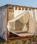Wooden pergola with white curtains on a beach, featuring a lounge bed inside, set against an ocean background.