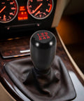 Upgrade Your Ride With This Sleek Kisa Angel 2 Piece Weighted Shift Knob Set For Manual Cars Featuring Ergonomic Grip, Anodized Finish, And Universal 8 10 12 Mm Adapter Kit For Smooth Gearing Action. Black aluminum manual shift knob with red 5-speed pattern in car interior