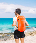 Man wearing 35L Swim Buoy Dry Bag Floating Backpack with Shoulder and Waist Straps for open water swimming on the beach, bright orange.