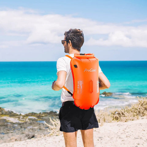 Man wearing 35L Swim Buoy Dry Bag Floating Backpack with Shoulder and Waist Straps for open water swimming on the beach, bright orange.