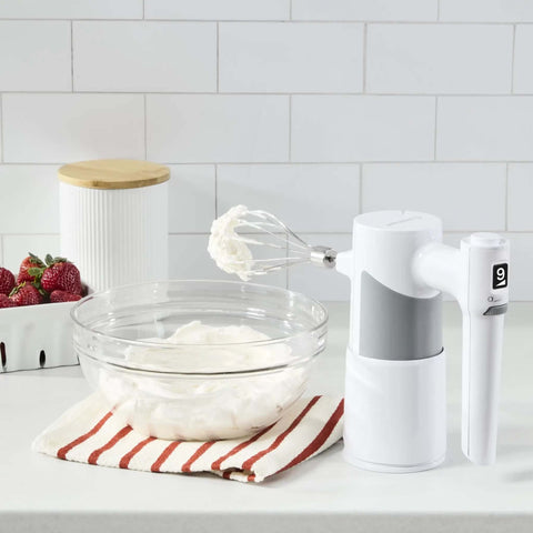 Lightweight Nine Speed Electric Hand Mixer With Grippy Ergonomic Handle And One Touch Digital Controls Built Different For Fast Whipping next to a bowl of whipped cream.