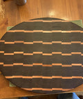 Wooden oval cutting board with dark and light wood pattern, handles on sides, displayed on wooden table.