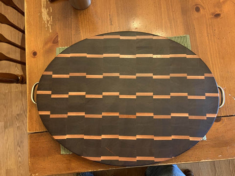 Wooden oval cutting board with dark and light wood pattern, handles on sides, displayed on wooden table.