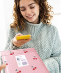Woman holding a phone and pink bubble mailer with cherry design; Pink Bubble Mailers 6x10 Inch 100 Pack Waterproof Self Seal Padded Envelopes