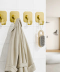 Heavy Duty Adhesive Wall Hooks In A Dope Four Pack, Stainless Steel Stick On Hangers With Badass Grip For Bathrooms And Kitchens, Waterproof And Super Fast To Put Up On Any Wall Or Door Without Drilling, Classic Gold Finish holding a coat on a tiled wall.