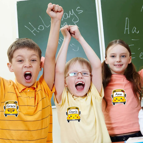 Children wearing school bus name tags in classroom with green chalkboards, showcasing 120 Pack School Bus Name Tags In 6 Cartoon Styles.