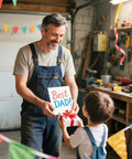 Man in workshop receiving 'Best Dad' card from child, surrounded by colorful decorations.
