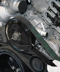 Pulley holder tool clamped on camshaft pulley in a car engine; black metal lever with timing belt and pulleys visible.