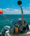 Boat throttle control levers on a sunny day, featuring metal handles with red and black ball grips against an ocean backdrop.