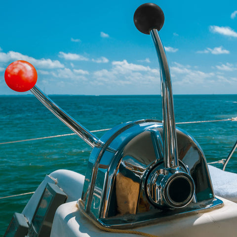 Boat throttle control levers on a sunny day, featuring metal handles with red and black ball grips against an ocean backdrop.