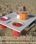 Portable Beach Table With Two Phone Slots, Lightweight Yet Sturdy And Durable, Must Have For Beach, Boat, Pool, Backyard, RV And Camping, Keeps Drinks And Phones Off The Sun With Built In Tray For Stash And Gear on sand with deli containers and blue cups.